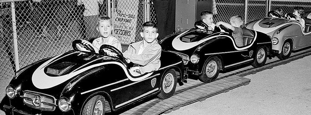 Elementary-school aged boys sit two-by-two in kid-sized amusement park race cars on an outdoor track in the evening. Men in 50s-60s garb stand behind a chain-link fence, watching over the kids. The photo is black and white.