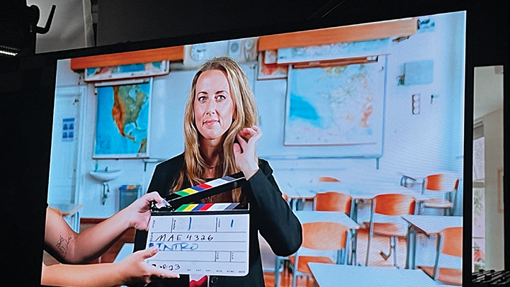 A young, blond woman looks to the camera as a director’s slate is held in front of her. A classroom setting with student desks is displayed behind her, on screen.