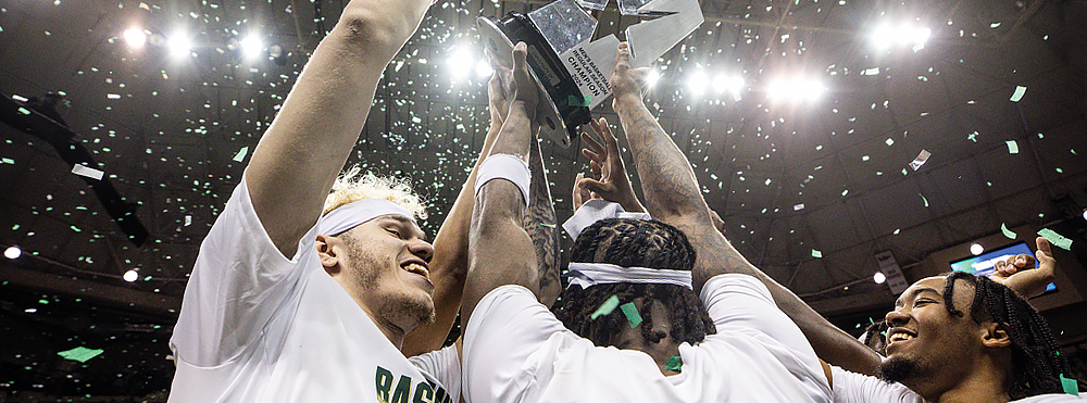Three USF men’s basketball players hold a large trophy overhead while bright lights and steaming confetti glow in the background. [Photo: USF Athletics]