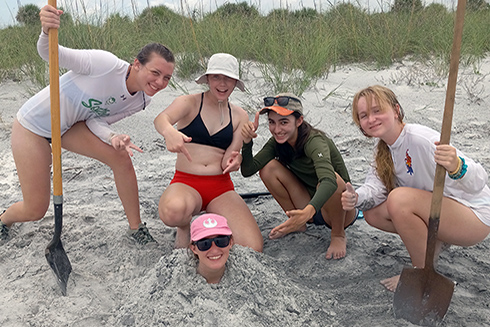 Campers Sarah, Hailey, Sarah, and Johnna bury science mentor Tiff using the pit they dug to observe beach sediment stratigraphy.