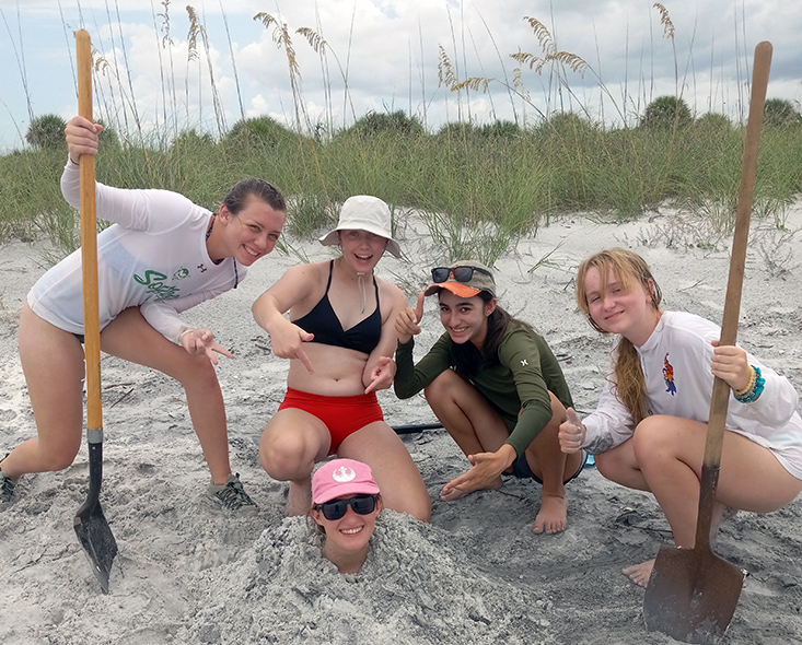 Campers Sarah, Hailey, Sarah, and Johnna bury science mentor Tiff using the pit they dug to observe beach sediment stratigraphy.