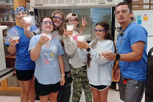 The lab rotation 2 group featuring OCG Fellow Shannon, campers Jocelyn, Arianna, Jenna, and Faustina, and Science Mentor Juan holding up their cool dry ice experiments in progress.