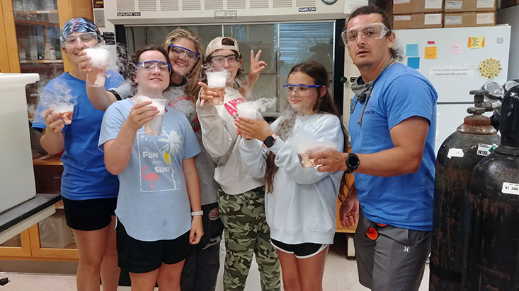 The lab rotation 2 group featuring OCG Fellow Shannon, campers Jocelyn, Arianna, Jenna, and Faustina, and Science Mentor Juan holding up their cool dry ice experiments in progress.
