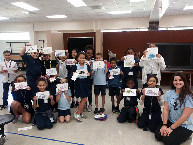 Makenzie Kerr with a 3rd grade class, holding up the fish coloring pages they got by deciphering which fish their egg was from. 