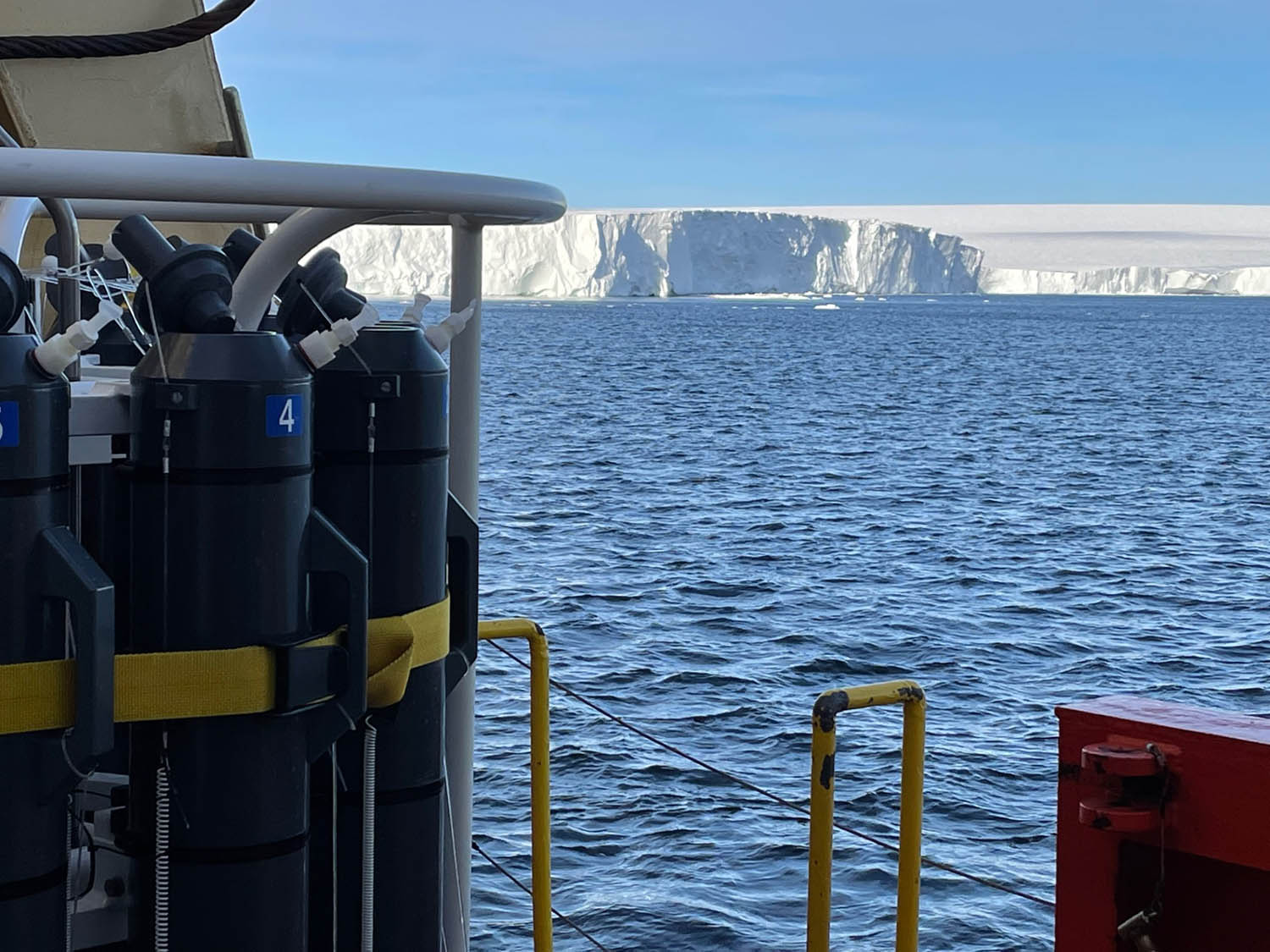 Sampling rosette with gray sampling bottles at left, the ship’s rail at lower right, and the face of the ice shelf in the background. Credit: Rob Sherrell