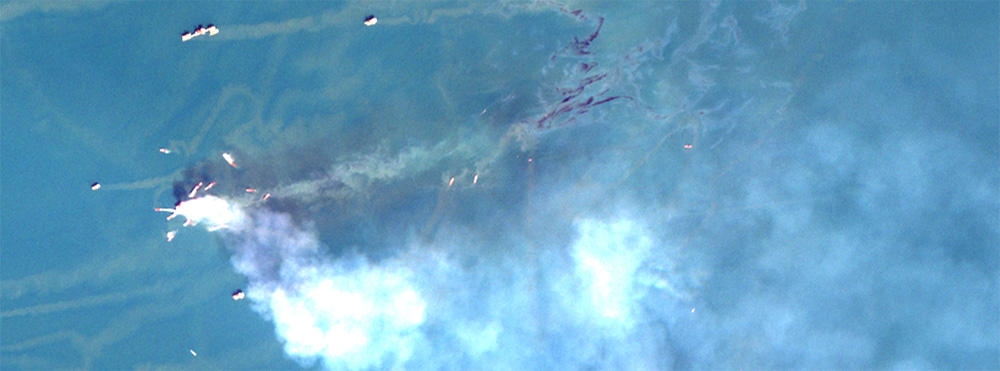 Oil spill from a fire-damaged platform, captured by the Sentinel-2 satellite of the European Space Agency. Fire smoke appears white, while oil slicks appear dark (crude and emulsified oil) or metallic color (oil sheen). Credit: Sentinel-2 data from Copernicus operated by the European Union Agency for the Space Programme in partnership with the European Space Agency. Image generated by Chuanmin Hu of University of South Florida.