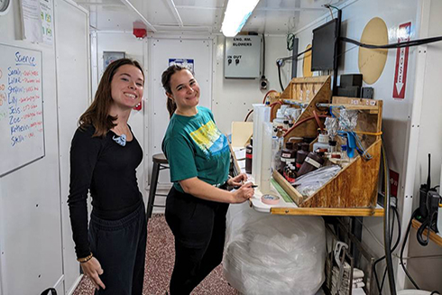 COMIT Interns, Zoe Brooker (left) and Katherin Abreus-Rodriguez (right), on the cruise next to the water sampling processing set-up. PHOTO CREDIT: Sarah Grasty.