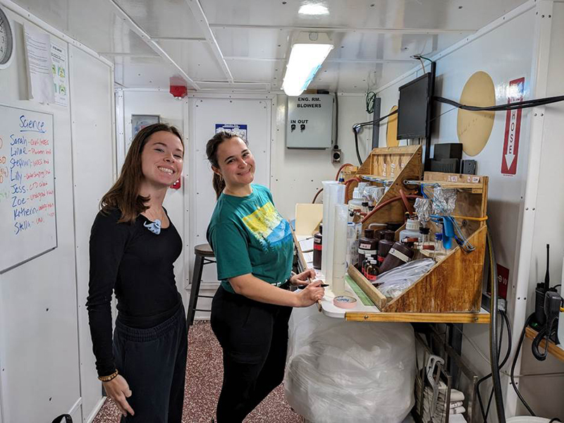 COMIT Interns, Zoe Brooker (left) and Katherin Abreus-Rodriguez (right), on the cruise next to the water sampling processing set-up. PHOTO CREDIT: Sarah Grasty.