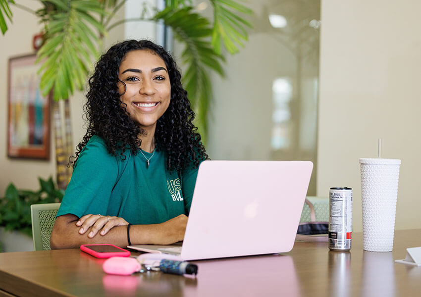 USF student smiles while sitting at a table with an open laptop.