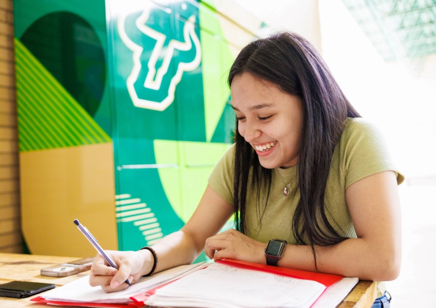 USF student smiles while sitting at a table and writing on paper.