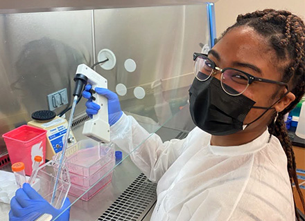 cason, wearing a white lab coat, looks over her shoulder as she uses a dropper to put liquid in a beaker