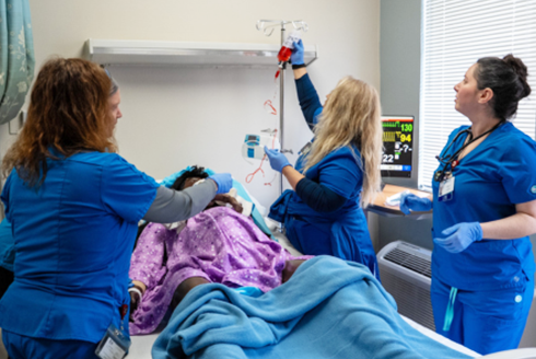 Three nurses stand over mannequin in hospital room