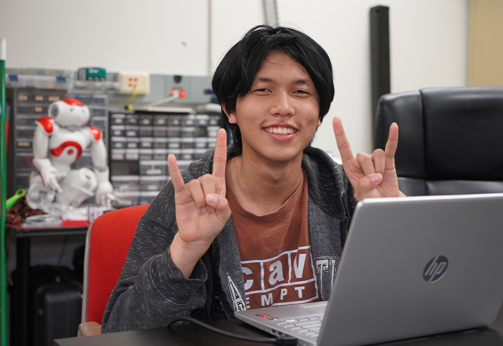 Rice Pham throwing up bulls sign behind his computer in robotics lab
