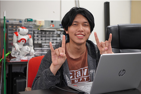 Rice Pham throwing up bulls sign behind his computer in robotics lab