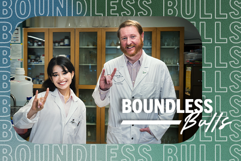 Claudia and Les standing in a lab, wearing white coats, smiling, and making the 