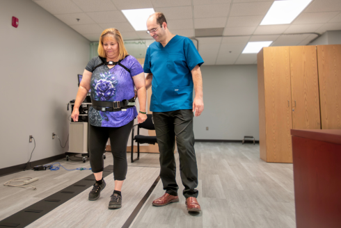 Patient walks with a device around waist on a designated pathway inside the clinic while guided by medical professional.