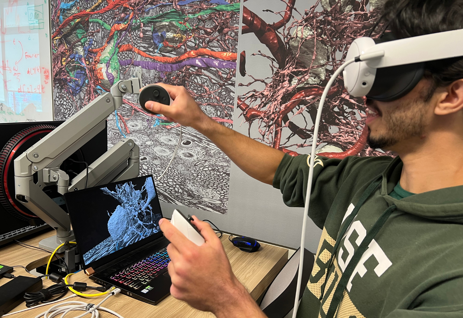 Student wearing VR goggles holds controllers in hands in front of computer with images of neurons