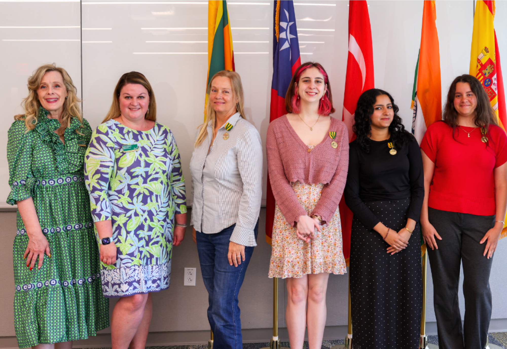 Kiki Caruson, Lauren Chambers, Elisabeth Parker, May Spinney, Afreen Khan, and Lizt Pena stand in front of their Fulbright country's flags in the Judy Genshaft Honors College.