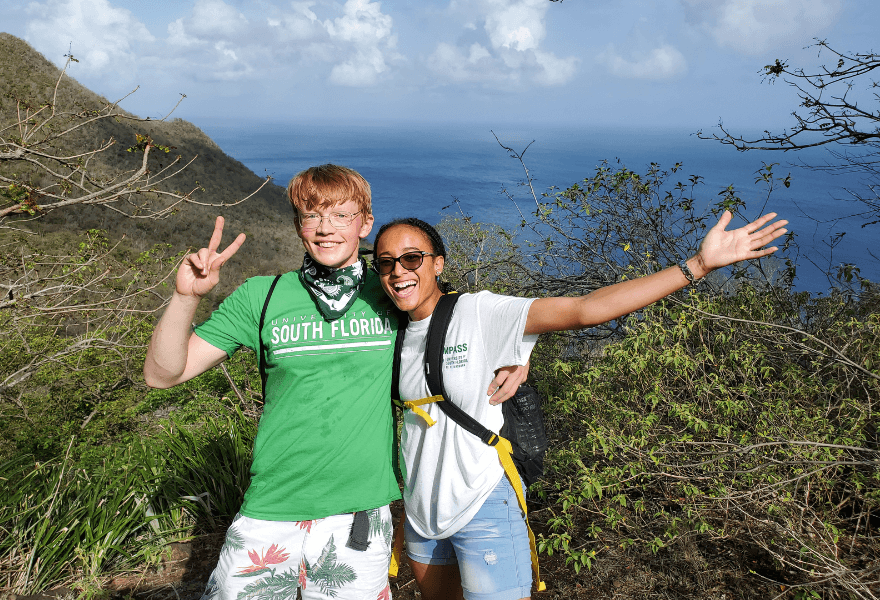 Two students in USF gear pose on a green cliff by the ocean