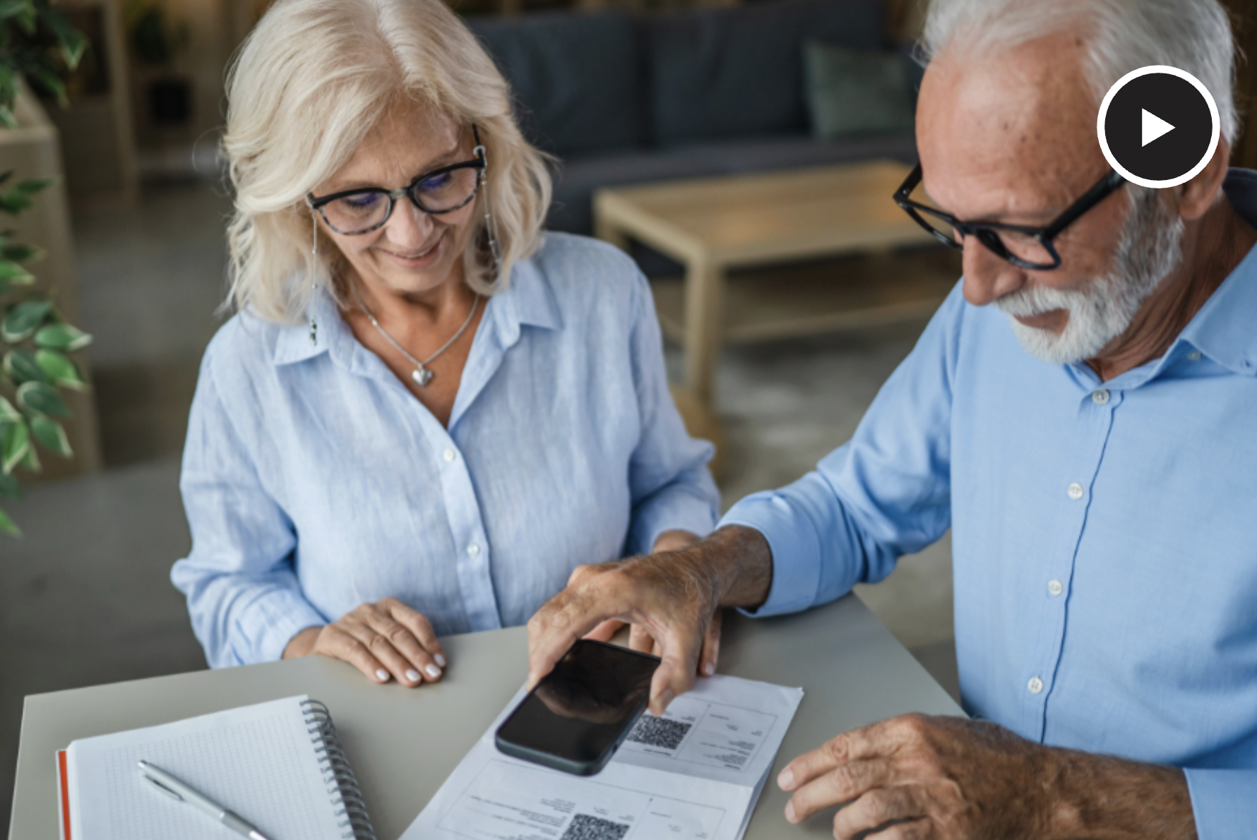 two older adults holding phone over QR code with play button to watch video