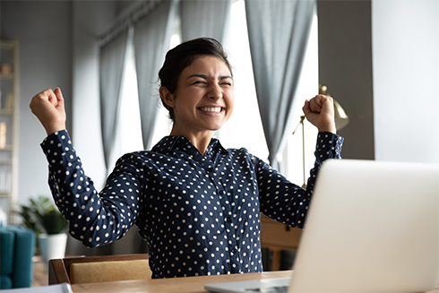 young woman with big smile and arms raised in celebration in front of her laptop like she just landed the job