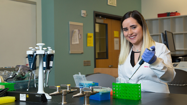 student Maria in white coat working in a research lab with pipette and test tubes