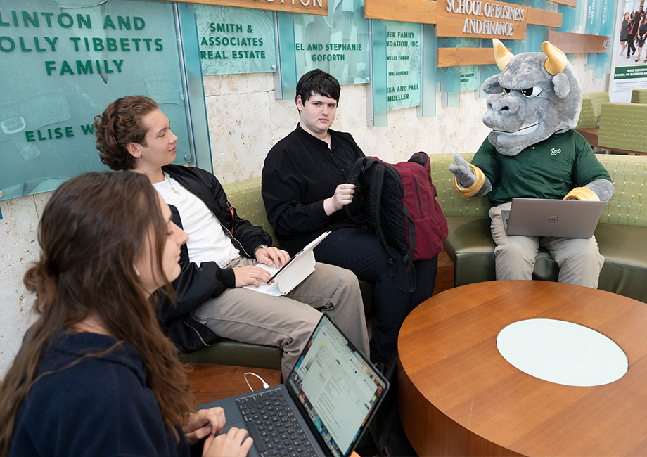 Rocky the Bull with a laptop on his lap, sitting next to three students