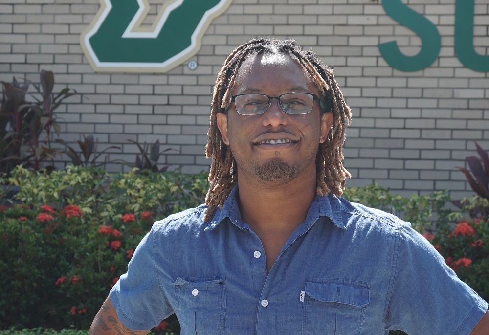 Dark-skinned male with dreadlocked hair and glasses standing in front of a USF sign wearing a demin shirt.