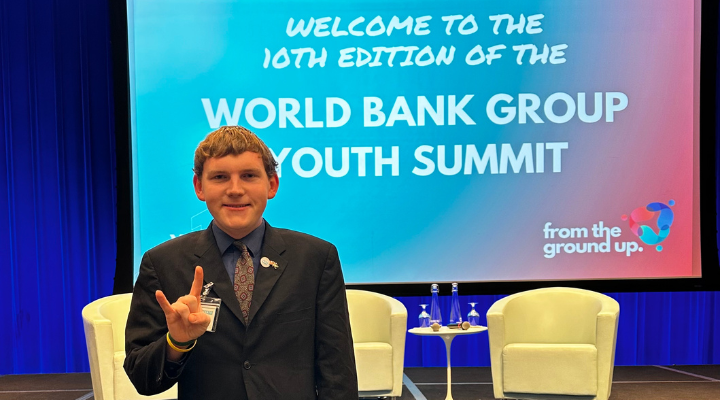 young white male standing in a dark suit on a stage infront of the welcome to the 10th edition of the World Bank Group Summit welcome sign
