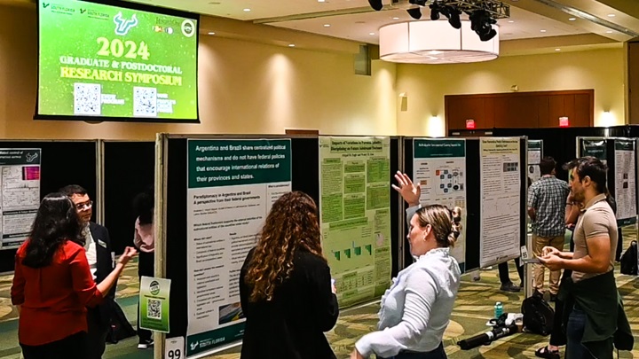 room view of scholars standing with their research posters and speaking with symposium guests