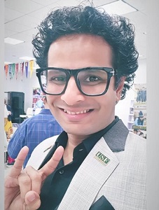 Indian alumnus, Ojas Rawal, smiling at the camera with his black curly hair, black glasses, dark shirt, and light grey suit jacket