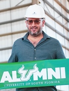head shot of USF alumnus Rene Jacir, a middle-aged Latin male wearing a hard hat and a drak green-blue shirt and holding a USF alumni banner
