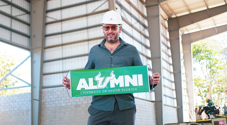 USG alumnus Rene Jacir - a middle-aged man in a hard hat wearing dark cargo pants and a button-down blue-green shirt in a hard hat - and holding a USF Alumni banner while standing in his company warehouse
