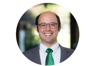 Headshot of a partially thinning gentleman with round tortise shell glasses wearing a charcoal suit jacket over a while shirt and a green tie