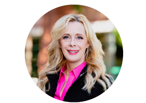 Headshot of a middle-aged blonde haired woman wearing a black suit jacket over a bright pink blouse.