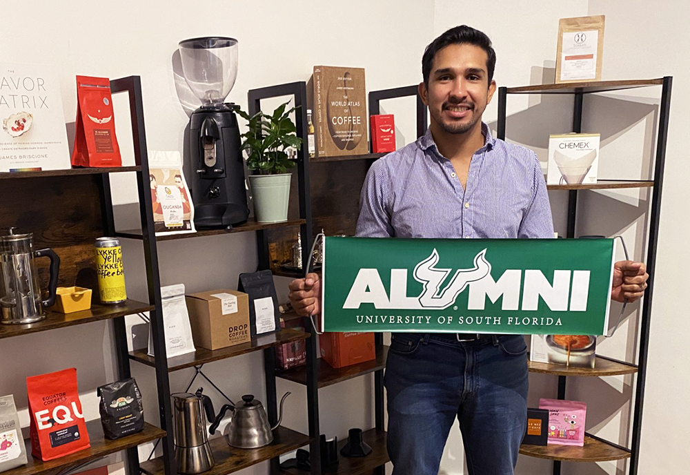 USF alumnus, Hernan Hererra standing in front of his coffee roasting equipment while holding a green and white USF alumni banner 