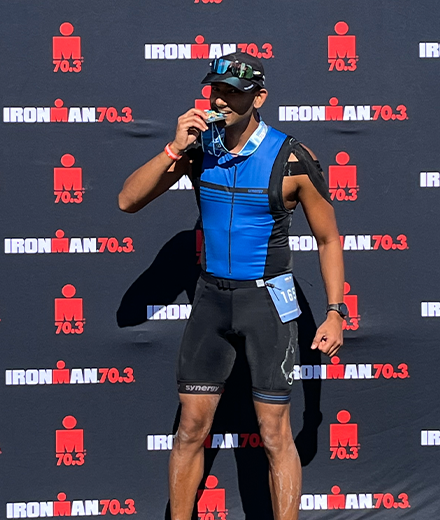 Sharma is standing before a backdrop that says "Ironman" and he is holding a gold medal and biting the end