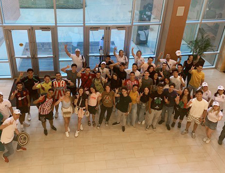 A group of people looking up at camera from the first floor, smiling and giving the bulls sign