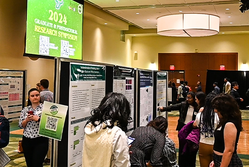 room view of scholars standing with their research posters and speaking with symposium guests