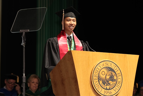 Vietnamese student, Kha Do stands at a podium on stage in regalia while giving a commencement speech