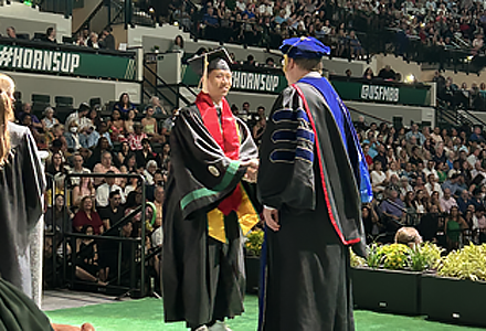 Vietnamese student, Kha So dressed in graduation regalia and shaking the hand at commencement of an administrator on the stage