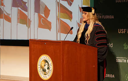 Dr. Kiki speaks at the Sash Ceremony at the podium. Behind her on a projector screen is an image of various flags for different countries represented at USF.