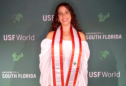 USF graduating student Hawi, posing for a photo with a USF green background. She is wearing a red and white sash representing Lebanon.