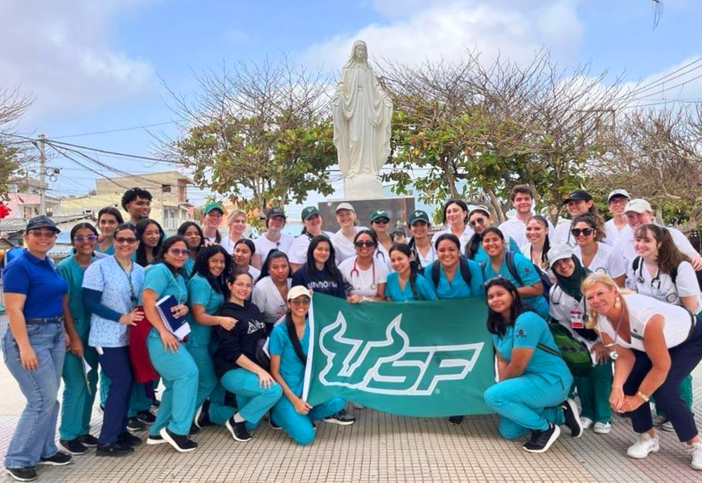 A group of USF nursing students in Colombia posing with a USF banner in front of a statue