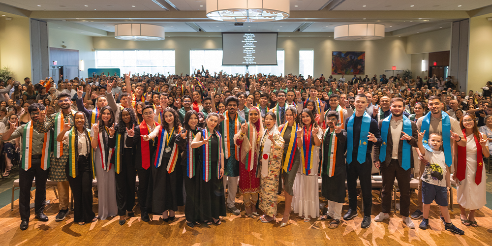 A large group of smiling graduating students who are holding up hands in the bulls symbol, all of the students wearing sashes that represent their home countries