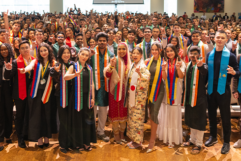 A large group of smiling graduating students who are holding up hands in the bulls symbol, all of the students wearing sashes that represent their home countries