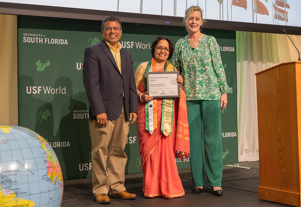 Provost, USF World Vice President, and Aruna, holding a certificate, are standing on stage smiling