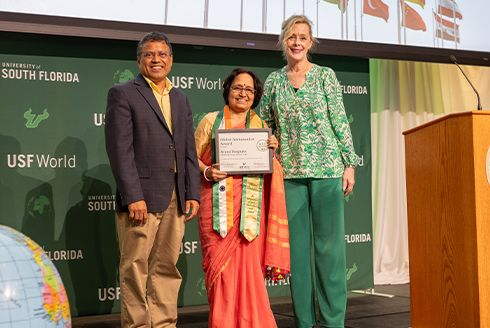 Provost, USF World Vice President, and Aruna, holding a certificate, are standing on stage smiling