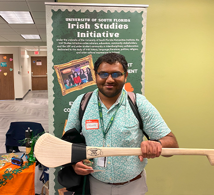 A person in a blue shirt holding a long paddle made of wood in front of a green sign that says "Irish Studies Initiative"