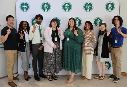 A group of people in front of a white backdrop with green logos, the group is holding up their hands in a bull sign